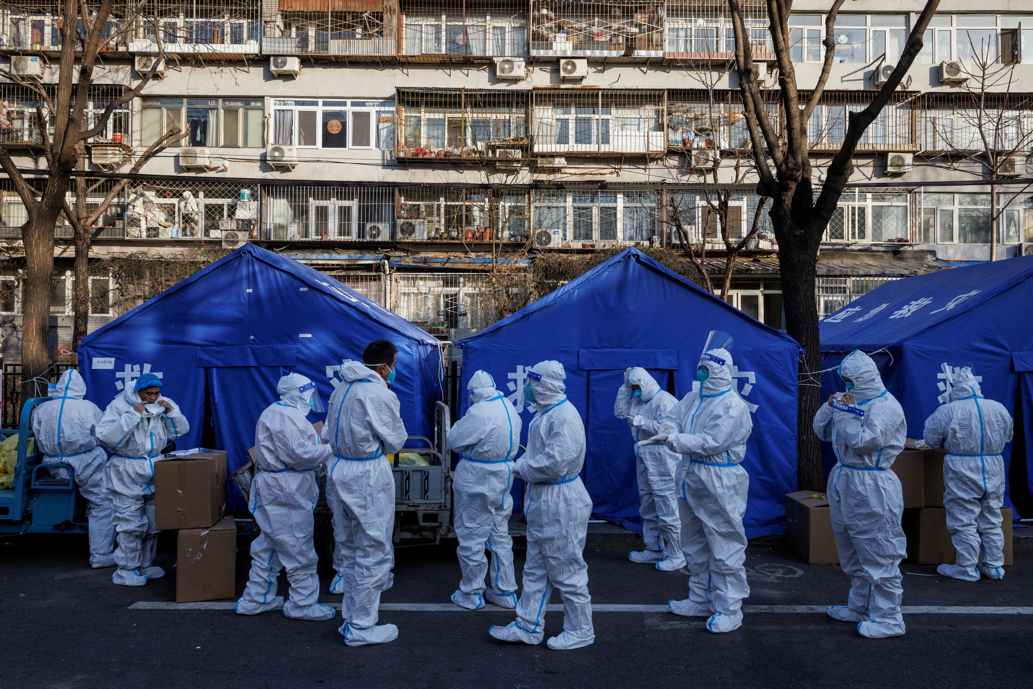 Pandemic workers in white hazmat suits gather in front of a block of flats in Beijing where people are under home quarantine. They are standing in front of blue tents and are about to begin their shift.